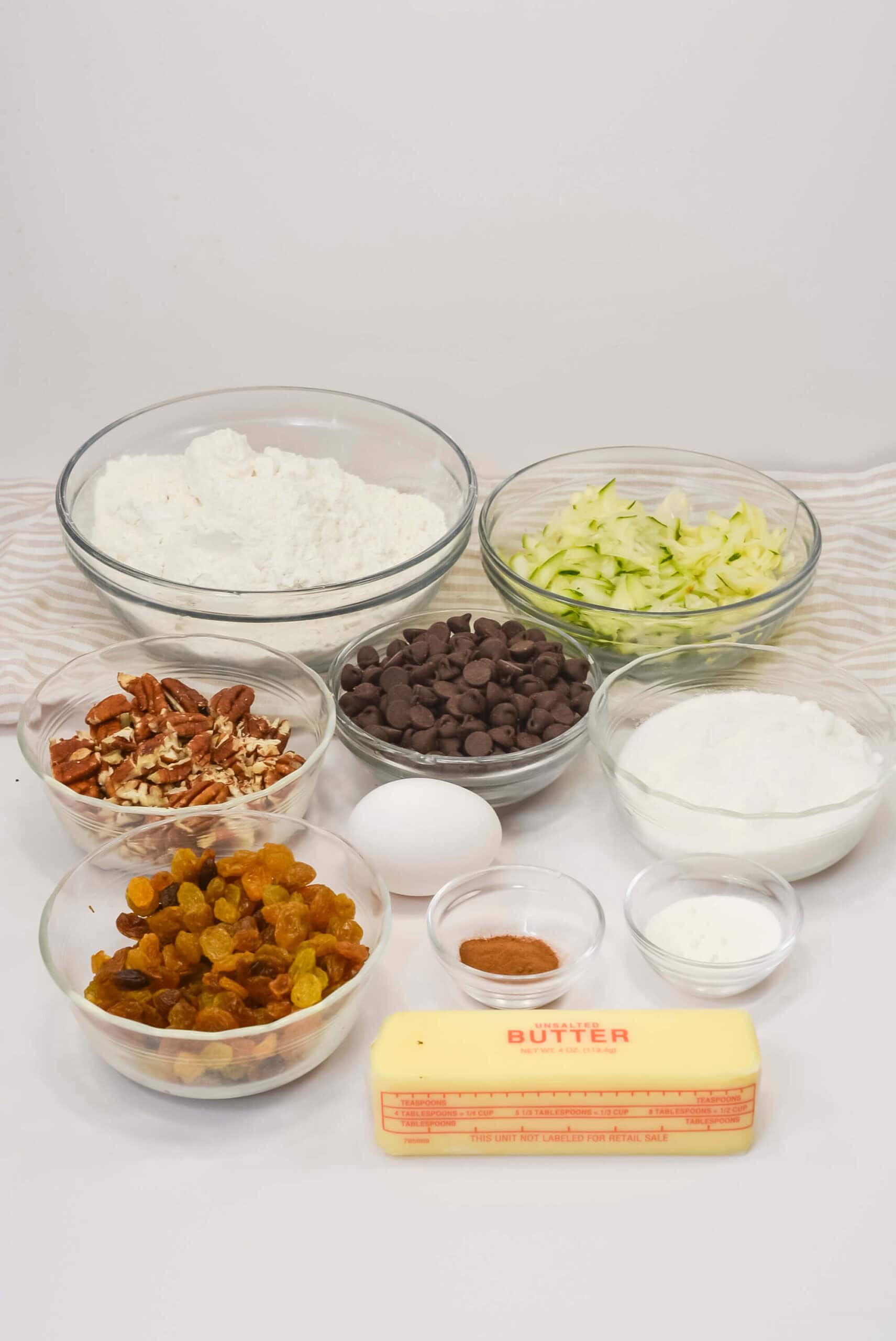 Various baking ingredients in bowls on a white surface are ready for baking including flour, shredded zucchini, chocolate chips, chopped pecans, raisins, an egg, sugar, cinnamon, baking powder, and a stick of butter.