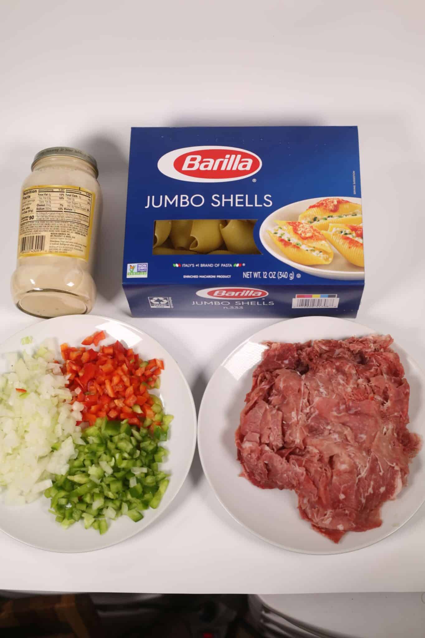 Box of Barilla jumbo shells, a jar of white sauce, chopped vegetables on a plate, and raw ground meat on another plate—perfect for creating Philly Cheesesteak Stuffed Shells.