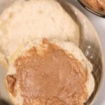 A biscuit, split in half and one side spread with Texas Roadhouse Butter, is placed on a beige plate. Nearby is a bowl containing more delightful cinnamon butter.