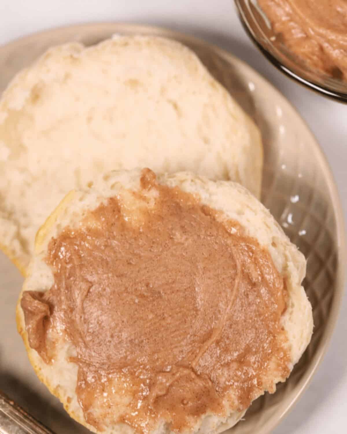 A biscuit, split in half and one side spread with Texas Roadhouse Butter, is placed on a beige plate. Nearby is a bowl containing more delightful cinnamon butter.