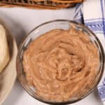 A small glass bowl filled with creamy, spread rests on a table near a basket with a blue and white checkered cloth.