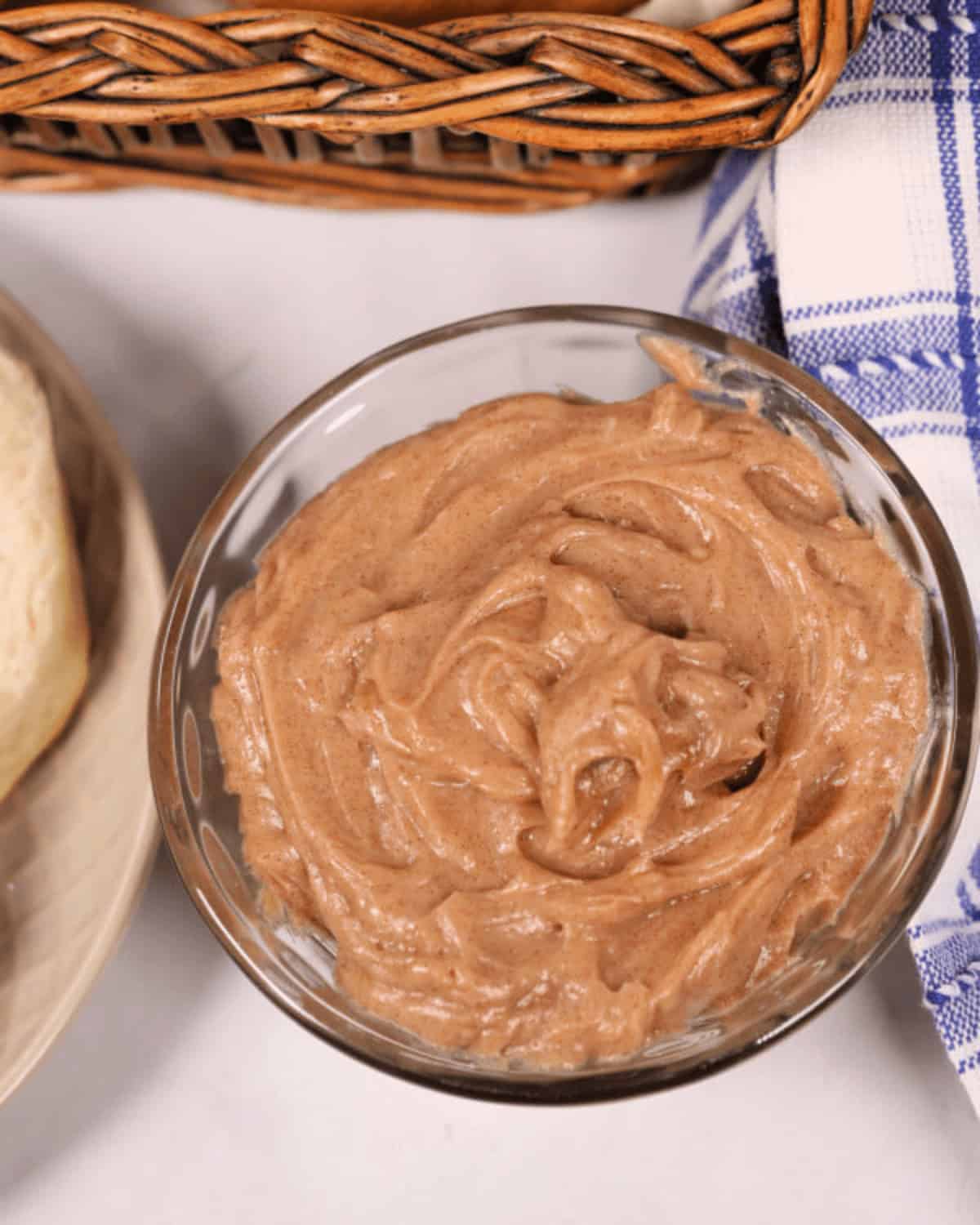 A small glass bowl filled with creamy, spread rests on a table near a basket with a blue and white checkered cloth.