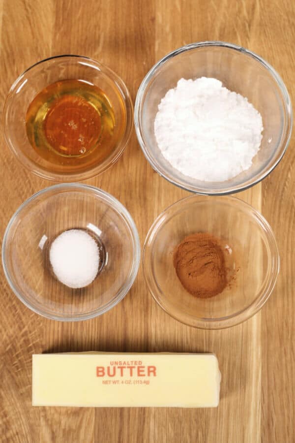 Five bowls on a wooden surface contain honey, powdered sugar, white sugar, cinnamon, and a stick of unsalted butter ready to be whipped into that delicious Texas Roadhouse Butter you're craving.