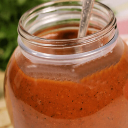 A jar of homemade tomato sauce with visible herbs and a spoon, on a table with a leafy green backdrop, ideal for chicken enchilada red sauce.