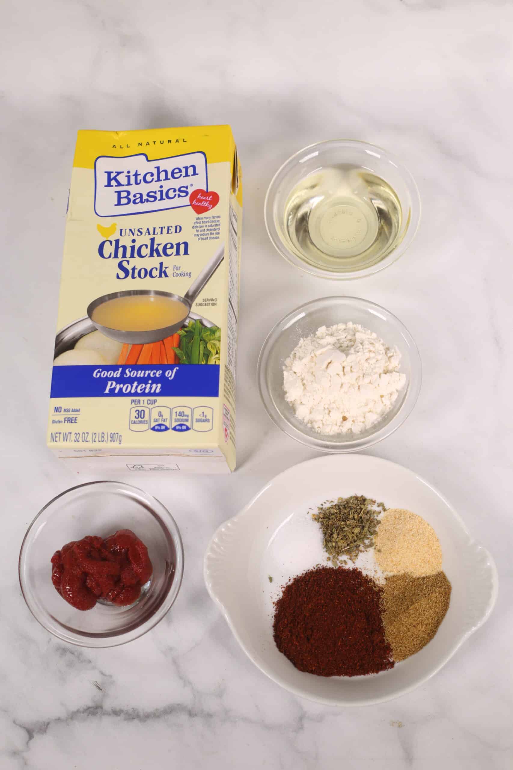 Ingredients for Chicken Enchilada Red Sauce on a marble countertop, including chicken stock, oil, flour, tomato paste, and assorted spices in various bowls.