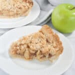 A slice of easy apple crumble pie on a white plate. Another slice of pie and a bowl of green apples are in the background.