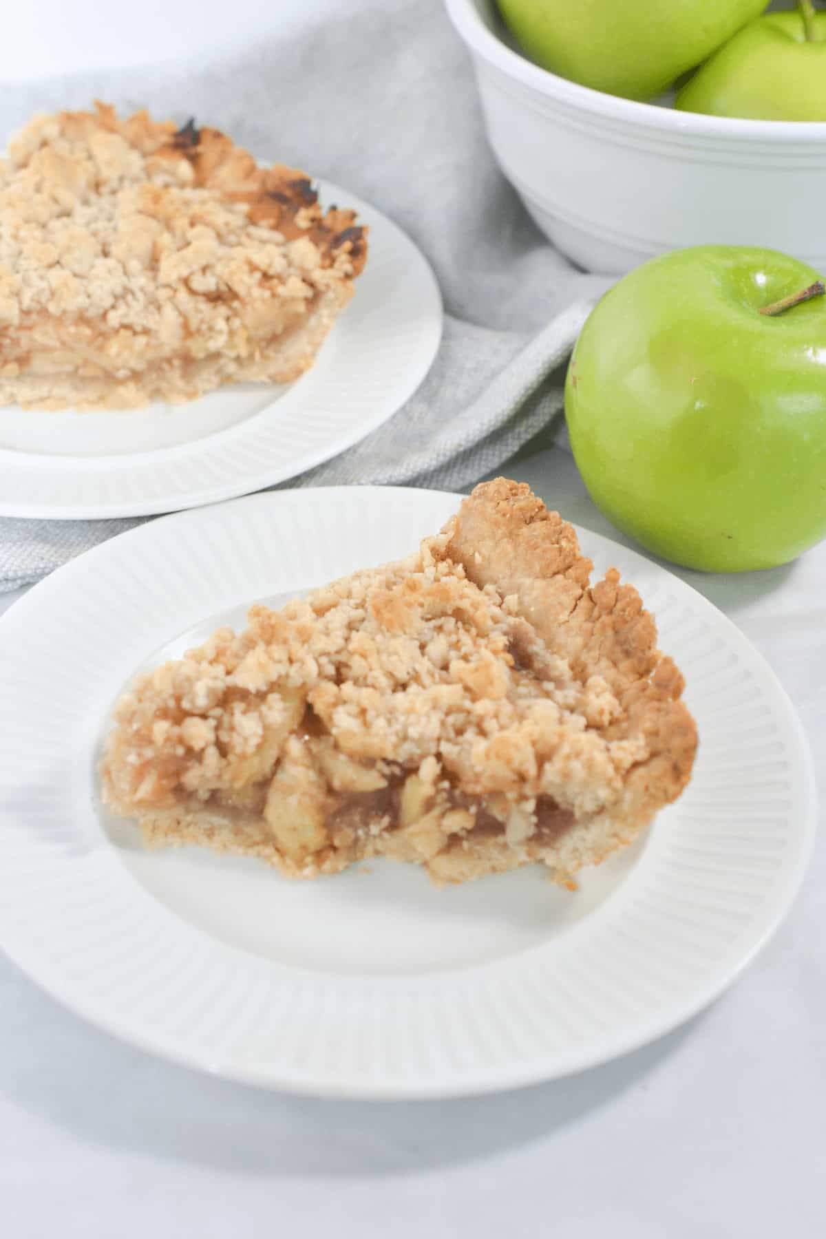 A slice of easy apple crumble pie on a white plate. Another slice of pie and a bowl of green apples are in the background.
