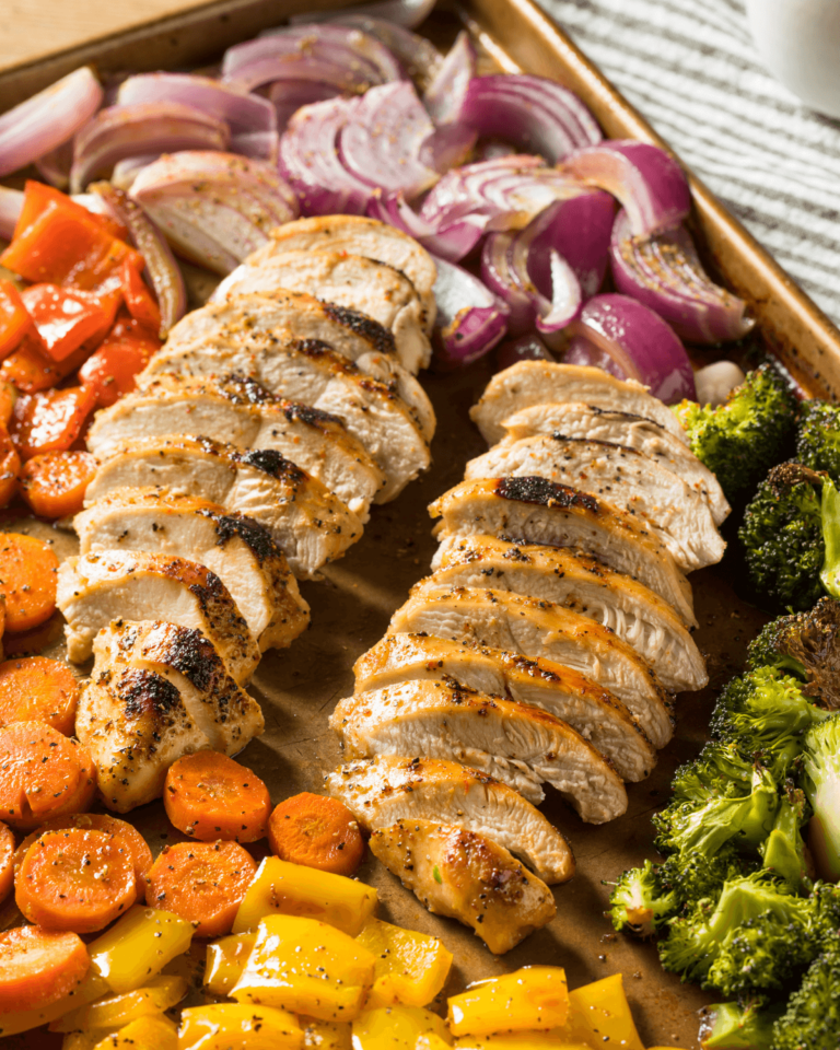 A balsamic chicken and vegetable sheet pan features slices of grilled chicken breast surrounded by roasted vegetables, including red onions, bell peppers, carrots, and broccoli.