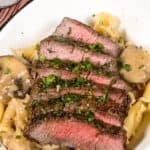 Sliced medium-rare steak served over pasta with a creamy mushroom and sherry cream sauce, garnished with chopped herbs.