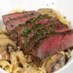 Sliced medium-rare steak served over pasta with savory mushroom and sherry cream sauce, garnished with chopped herbs in a white bowl.