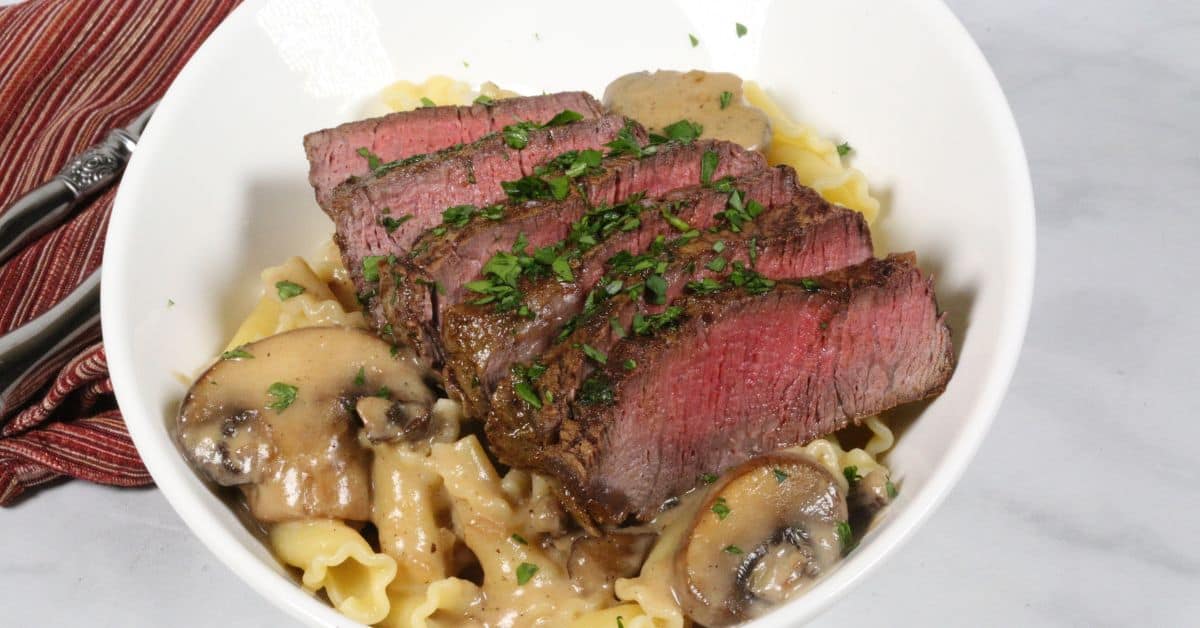 Sliced medium-rare steak served over pasta with savory mushroom and sherry cream sauce, garnished with chopped herbs in a white bowl.