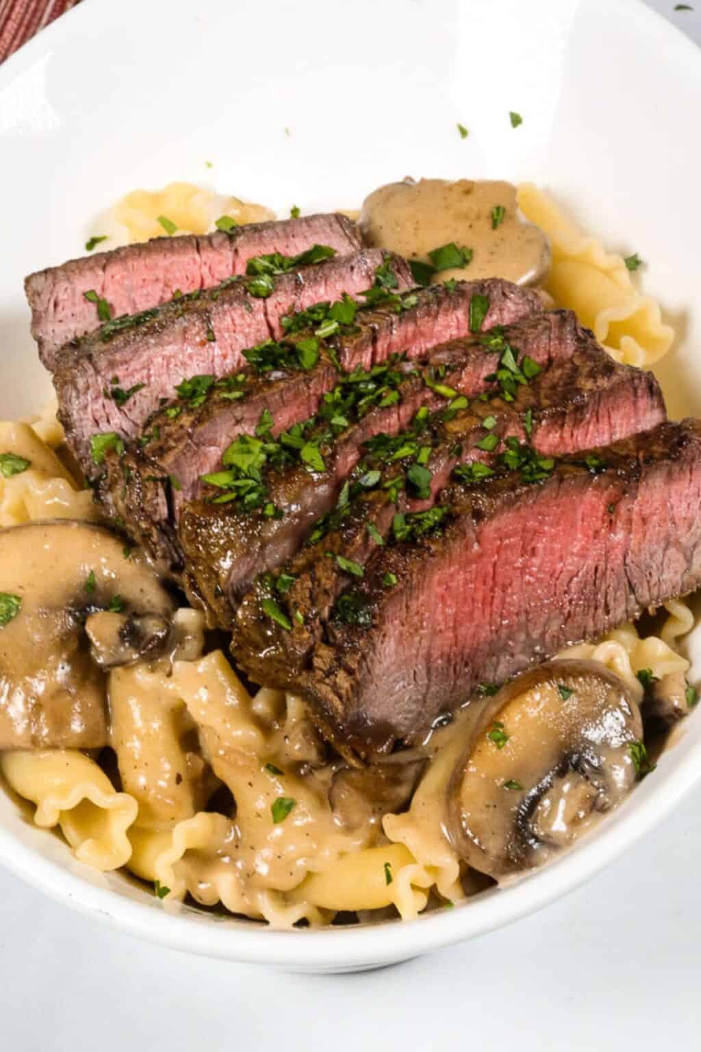 Sliced medium-rare steak served over pasta with a rich sherry cream sauce and mushrooms, garnished with chopped parsley in a white bowl.