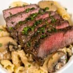 Sliced medium-rare steak served over pasta with a rich sherry cream sauce and mushrooms, garnished with chopped parsley in a white bowl.