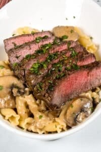 Sliced medium-rare steak served over pasta with a rich sherry cream sauce and mushrooms, garnished with chopped parsley in a white bowl.