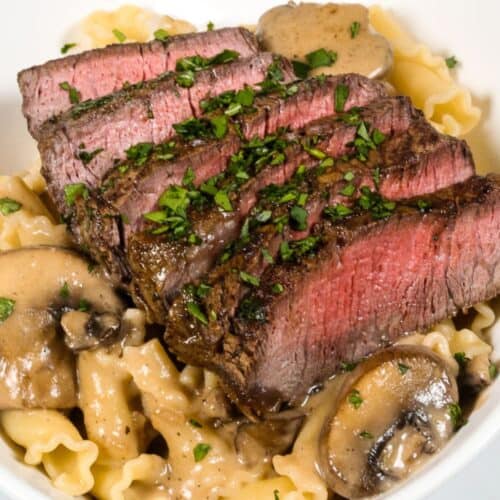 Sliced medium-rare steak served over pasta with a rich sherry cream sauce and mushrooms, garnished with chopped parsley in a white bowl.