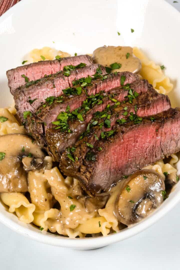 Sliced medium-rare steak served over pasta with a rich sherry cream sauce and mushrooms, garnished with chopped parsley in a white bowl.