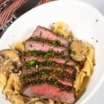 Sliced steak served over pasta with mushrooms garnished with chopped herbs in a white bowl; fork, knife, and red-striped napkin beside.