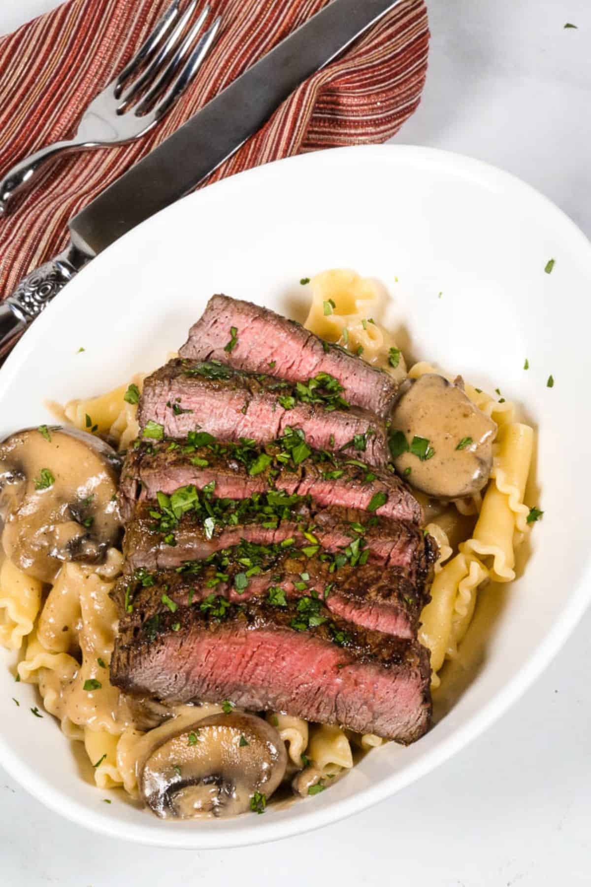 Sliced steak served over pasta with mushrooms  garnished with chopped herbs in a white bowl; fork, knife, and red-striped napkin beside.