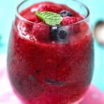 A glass filled with a vibrant slush, topped with fresh berries and a mint leaf, sits on a pink cloth against a blue background.