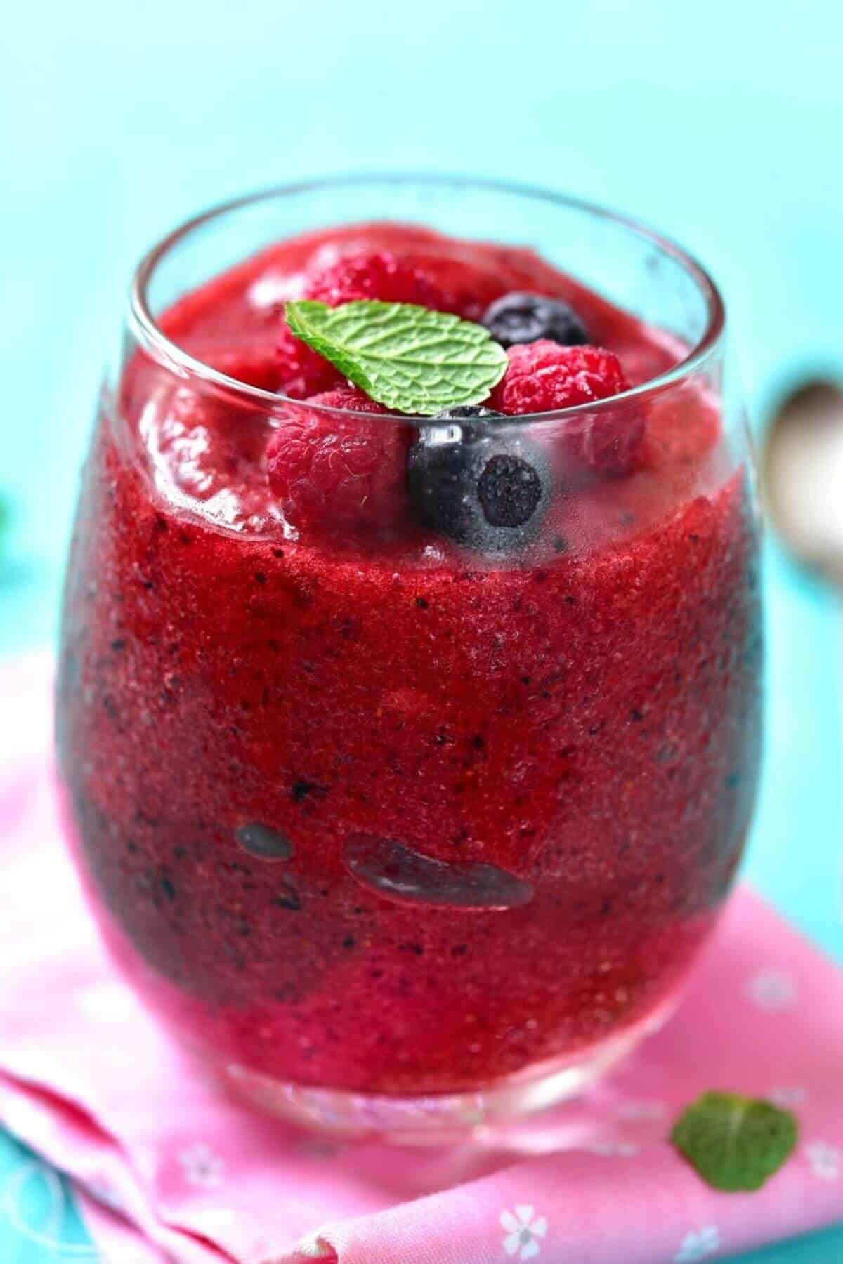 A glass filled with a vibrant slush, topped with fresh berries and a mint leaf, sits on a pink cloth against a blue background.