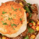 A close-up of a skillet beef pot pie topped with a golden-brown biscuit and garnished with fresh herbs. The stew includes pieces of beef, corn, carrots, and green beans.