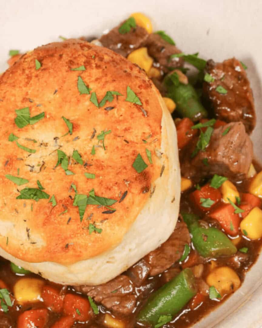 A close-up of a skillet beef pot pie topped with a golden-brown biscuit and garnished with fresh herbs. The stew includes pieces of beef, corn, carrots, and green beans.