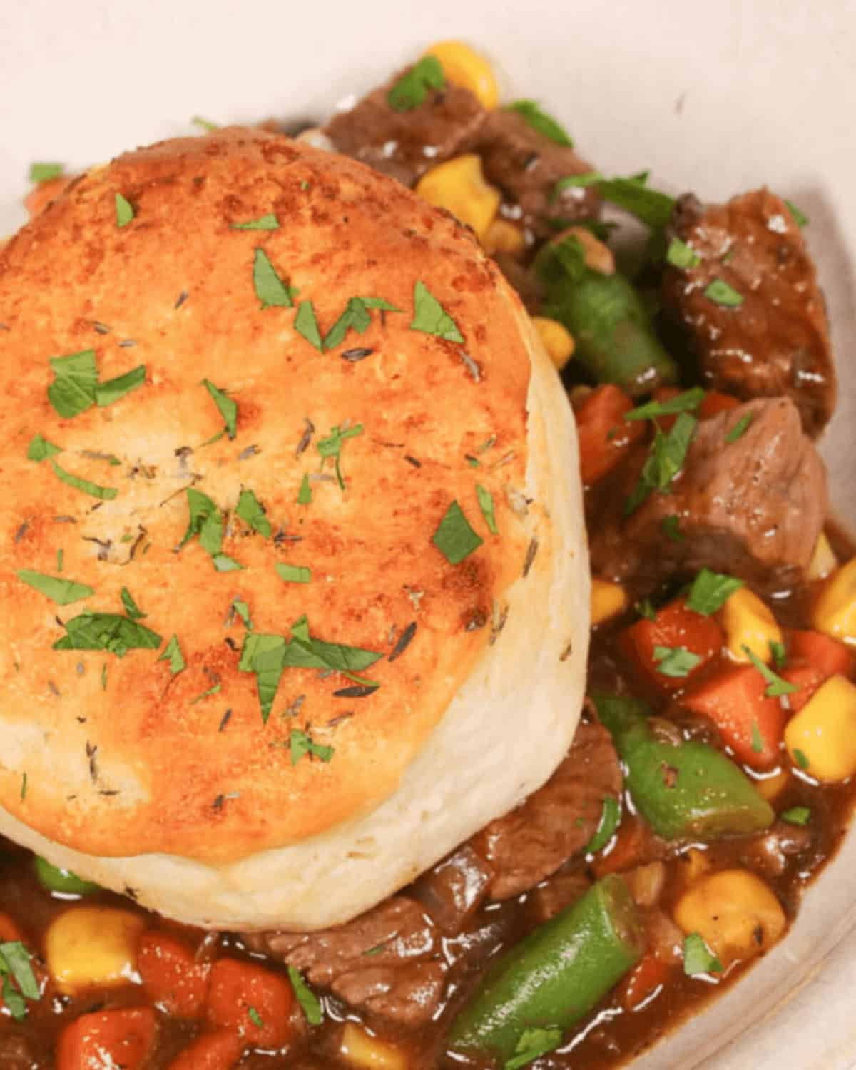 A close-up of a skillet beef pot pie topped with a golden-brown biscuit and garnished with fresh herbs. The stew includes pieces of beef, corn, carrots, and green beans.