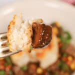 A fork holding a piece of biscuit and a chunk of meat with gravy, against a blurred background of a skillet beef pot pie.
