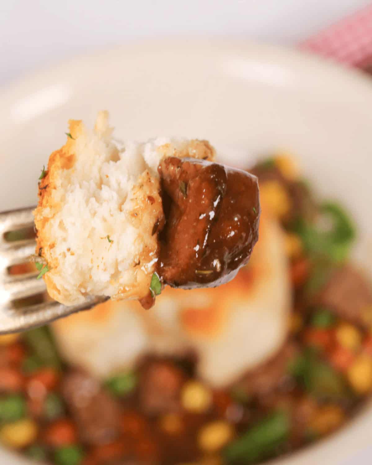 A fork holding a piece of biscuit and a chunk of meat with gravy, against a blurred background of a skillet beef pot pie.