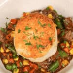 A bowl of with corn, green beans, peas, and carrots, topped with a golden biscuit reminiscent of skillet beef pot pie, garnished with chopped herbs.