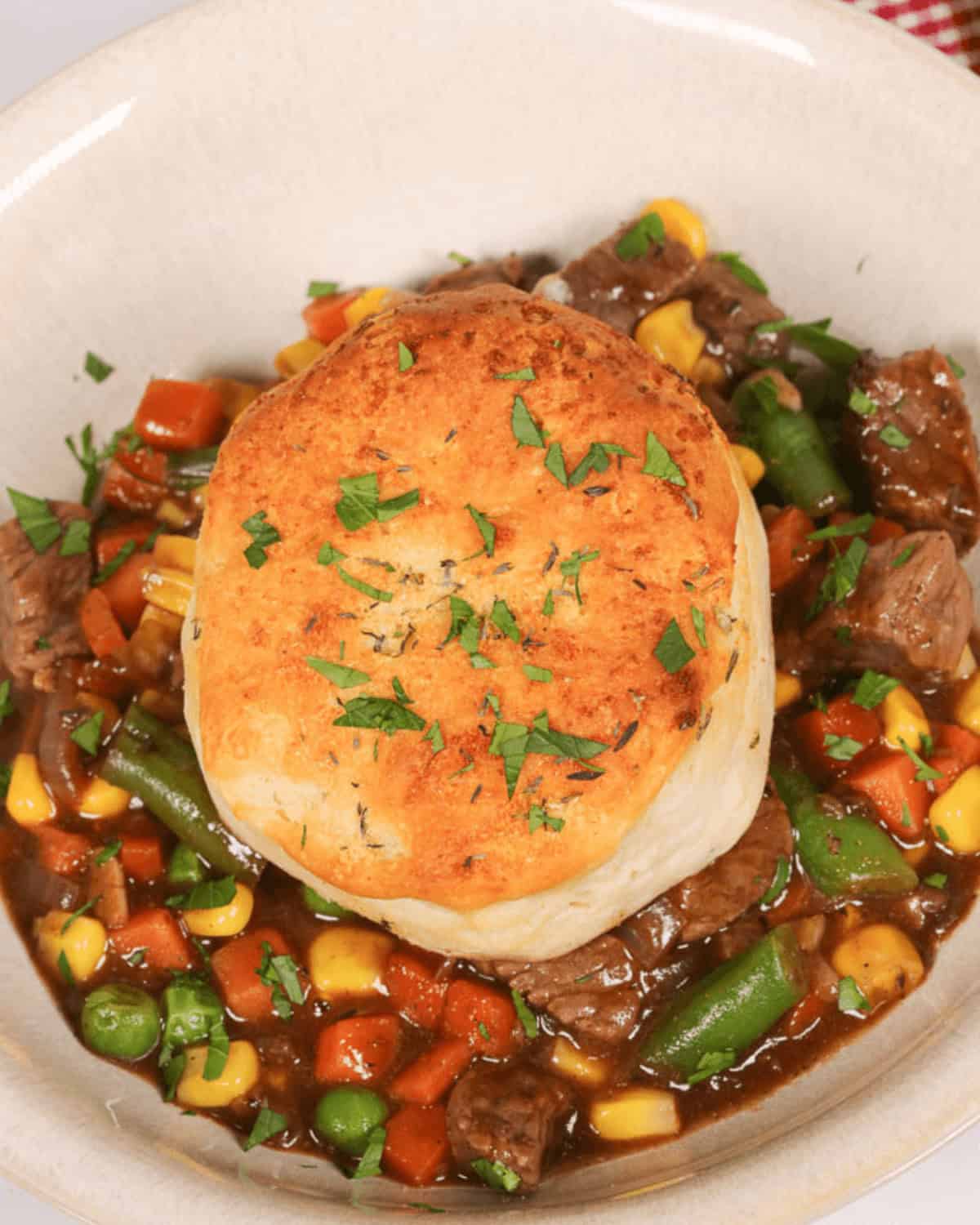 A bowl of with corn, green beans, peas, and carrots, topped with a golden biscuit reminiscent of skillet beef pot pie, garnished with chopped herbs.
