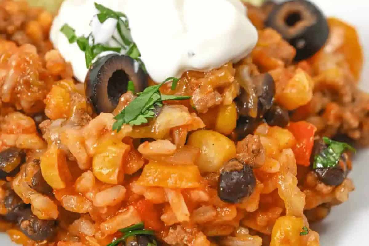 Close-up of a taco casserole with ground meat, black beans, corn, rice, sliced olives, and diced tomatoes, topped with fresh cilantro and a dollop of sour cream—perfect Cinco de Mayo food.