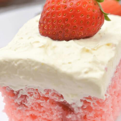 A slice of Strawberry Poke Cake topped with a thick layer of whipped cream and a whole strawberry placed on top, served on a white plate.