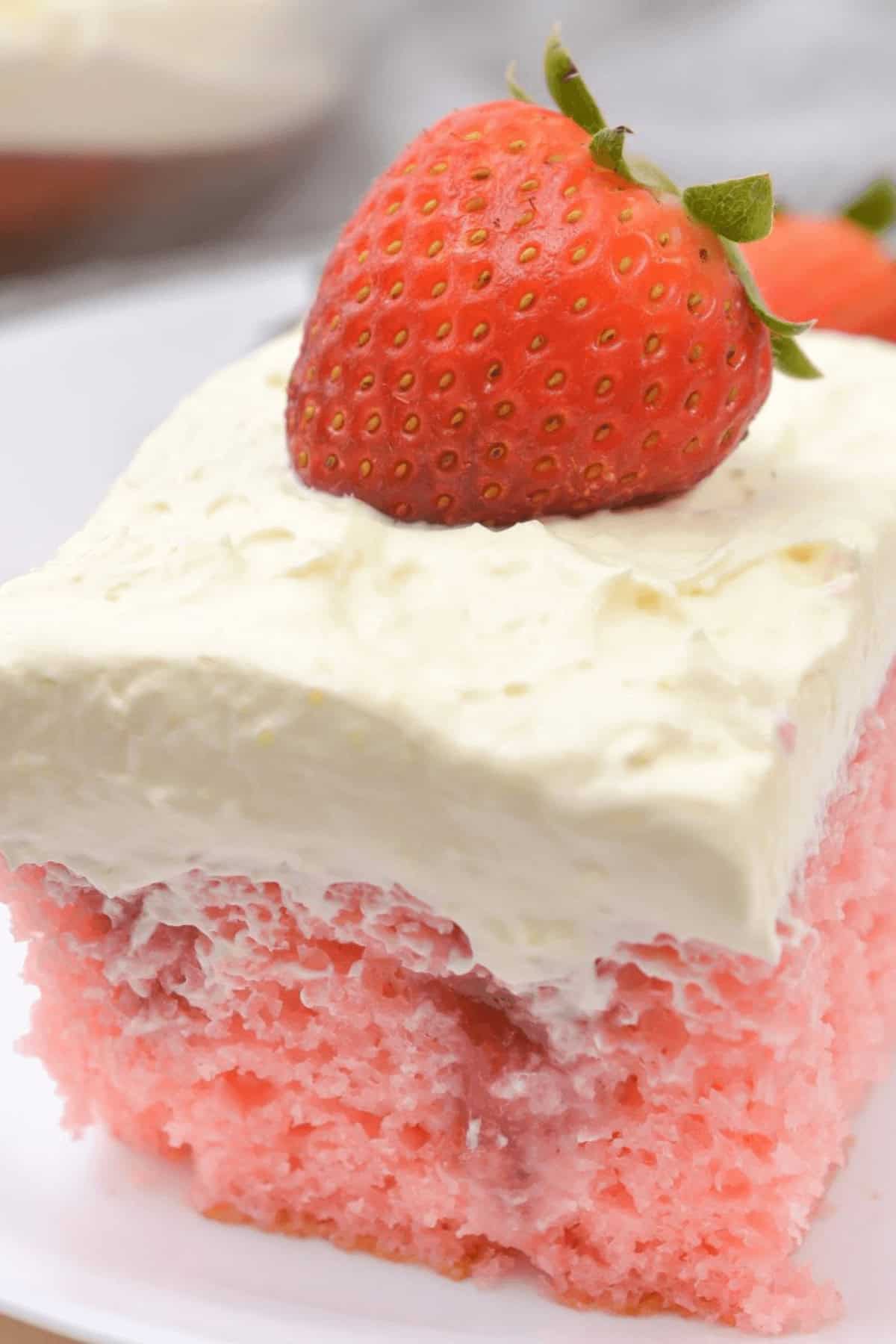 A slice of Strawberry Poke Cake topped with a thick layer of whipped cream and a whole strawberry placed on top, served on a white plate.
