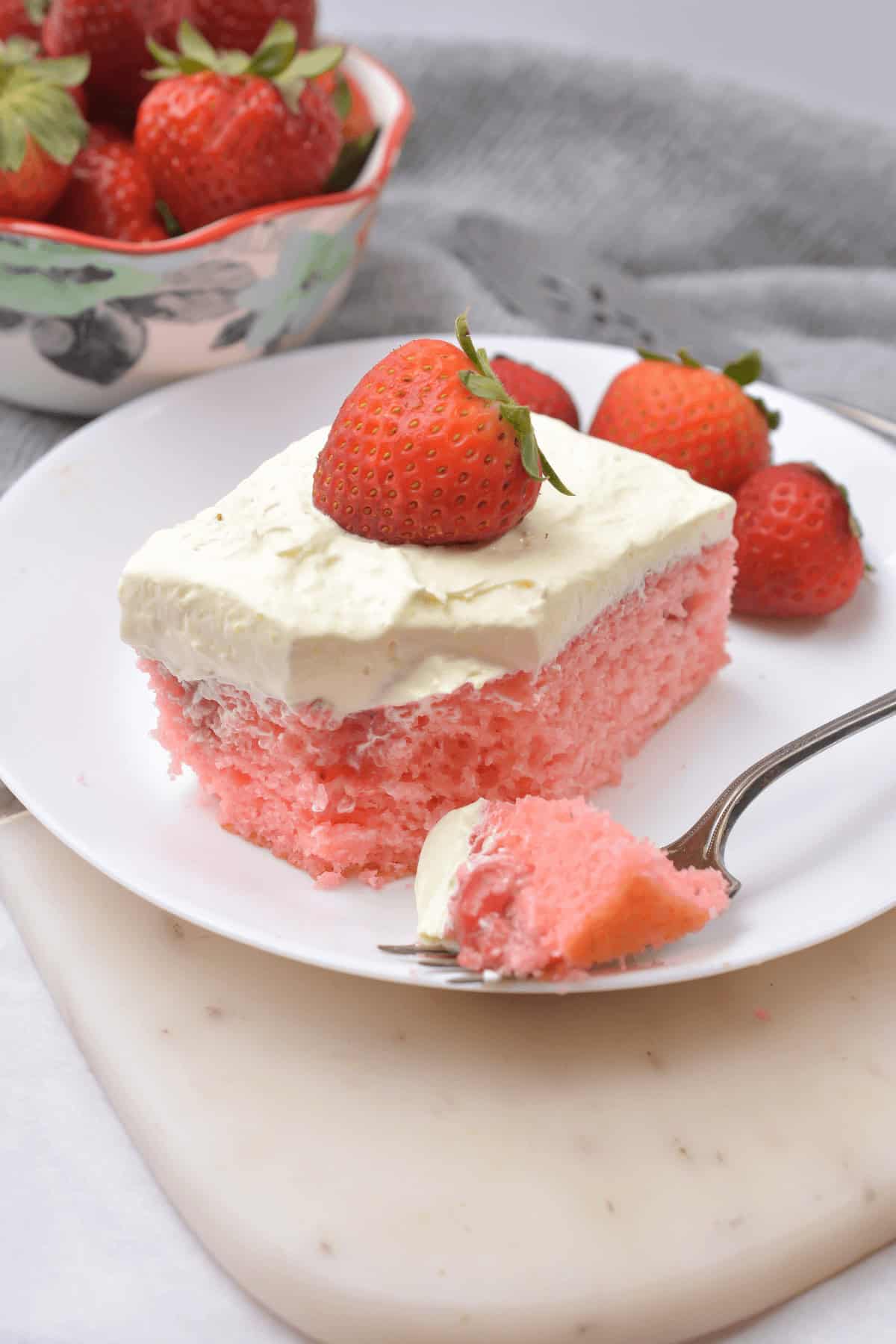 A slice of pink Strawberry Poke Cake with white frosting topped by a strawberry on a white plate with a forkful of cake. A bowl of strawberries is in the background.