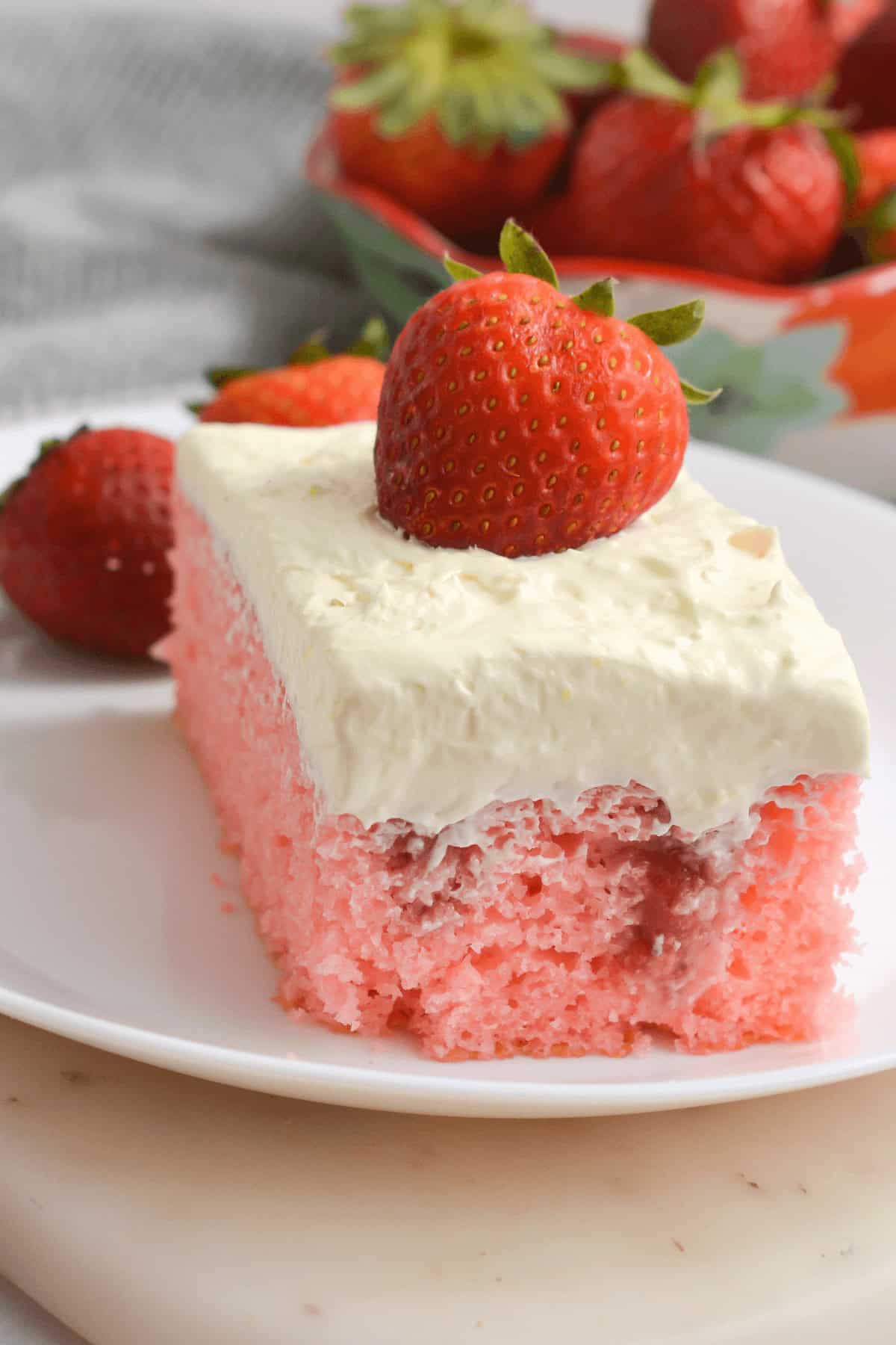 A slice of pink Strawberry Poke Cake topped with white frosting and a whole strawberry sits on a white plate. A bowl of strawberries is in the background.