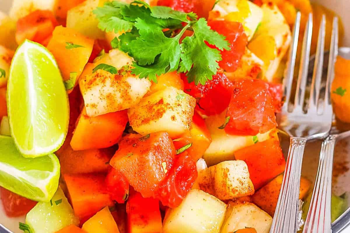 A colorful fruit salad with diced watermelon, melon, and papaya, garnished with cilantro—perfect as a refreshing Cinco de Mayo food. Lime wedges and two forks are placed beside the salad.