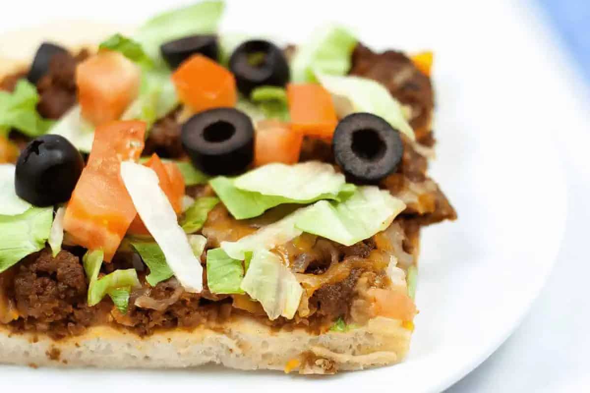 A festive square slice of taco pizza, perfect for Cinco de Mayo, features ground beef, melted cheese, shredded lettuce, diced tomatoes, and sliced black olives on a white plate.