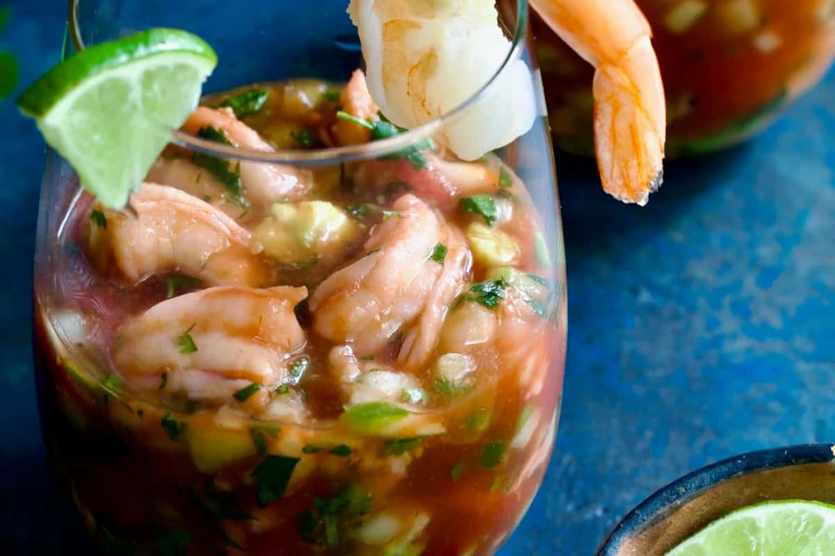 A glass filled with shrimp cocktail, a festive Cinco de Mayo food, is garnished with lime and herbs, set on a vibrant blue surface.