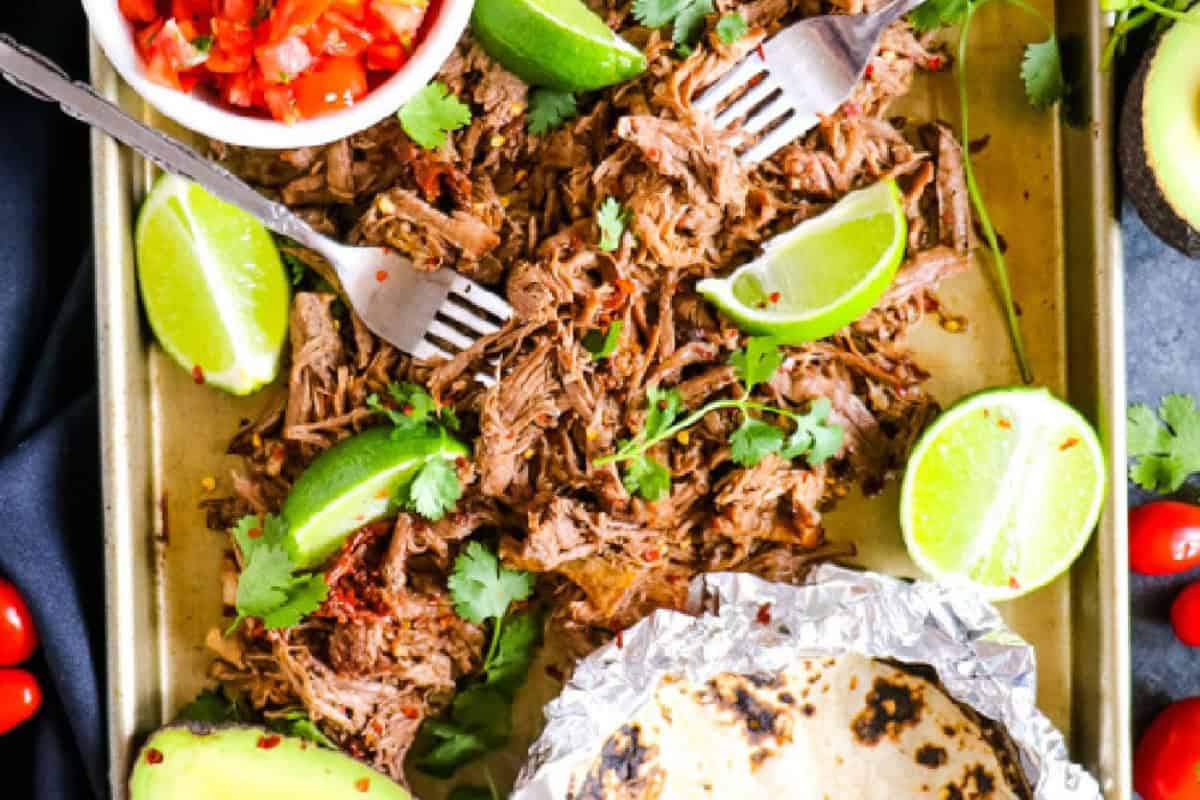 A tray of shredded beef garnished with lime wedges and cilantro, perfect for Cinco de Mayo festivities. On the side, a bowl of chopped tomatoes and soft tortillas wrapped in foil await. Creamy avocado halves complete this festive spread.