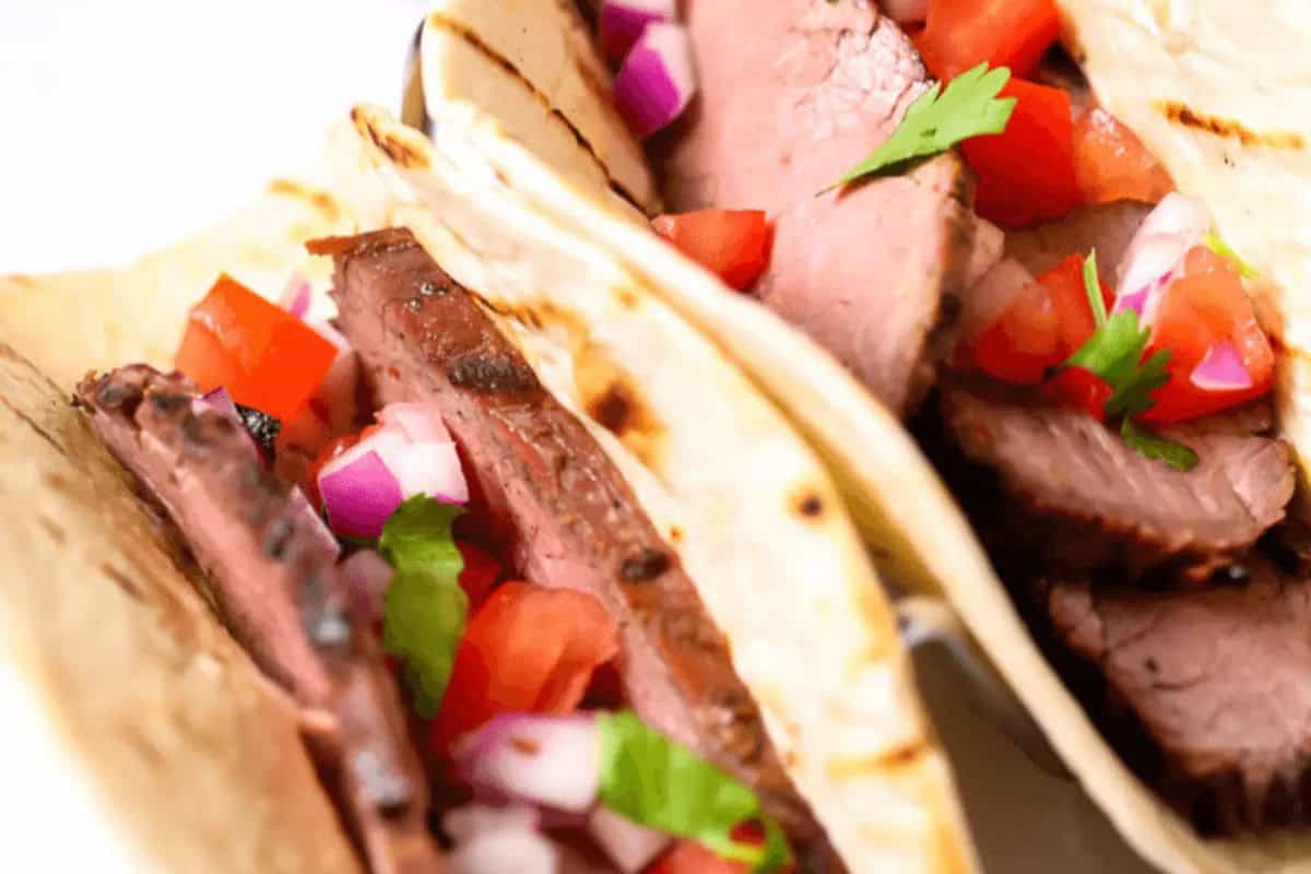 Close-up of two grilled beef tacos, a classic Cinco de Mayo food, filled with sliced beef, diced tomatoes, red onions, and cilantro on soft tortillas.