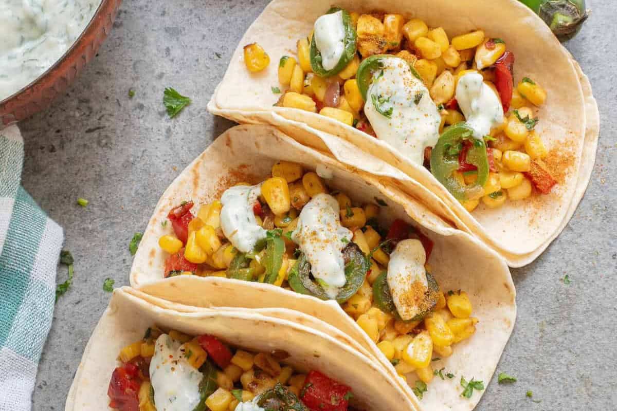 A trio of tacos brimming with corn, jalapeños, and diced red peppers, crowned with a creamy sauce—a perfect Cinco de Mayo food delight—rests invitingly on a gray surface.
