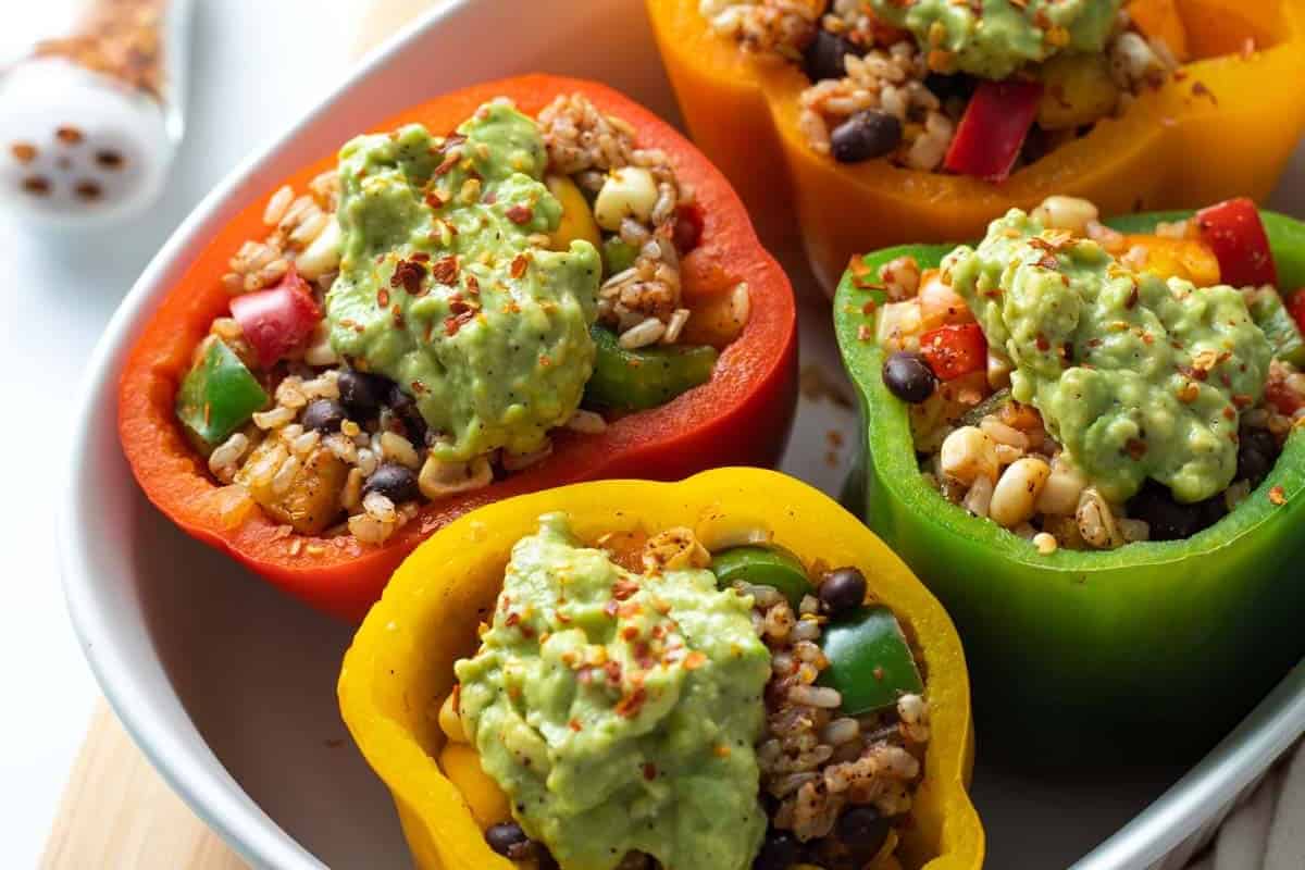 Four halved bell peppers filled with rice, beans, corn, and vegetables, topped with guacamole, sit in a white dish—a vibrant Cinco de Mayo food delight.