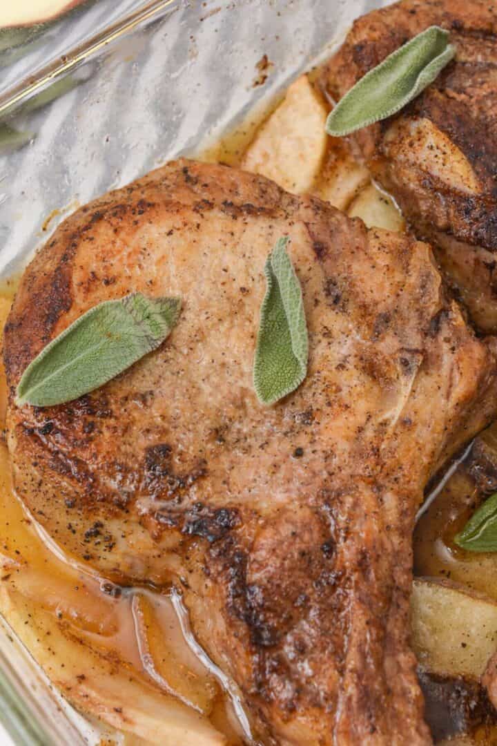 Close-up of pan-seared pork chops in a dish, garnished with fresh sage leaves, with visible juices at the bottom.