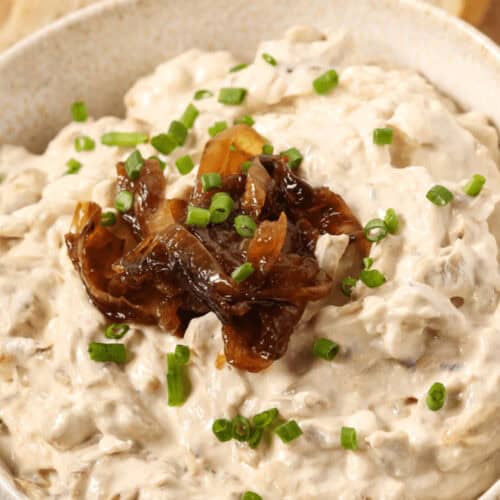 A bowl of rich caramelized onion dip is topped with chopped chives, with sliced bread in the background.