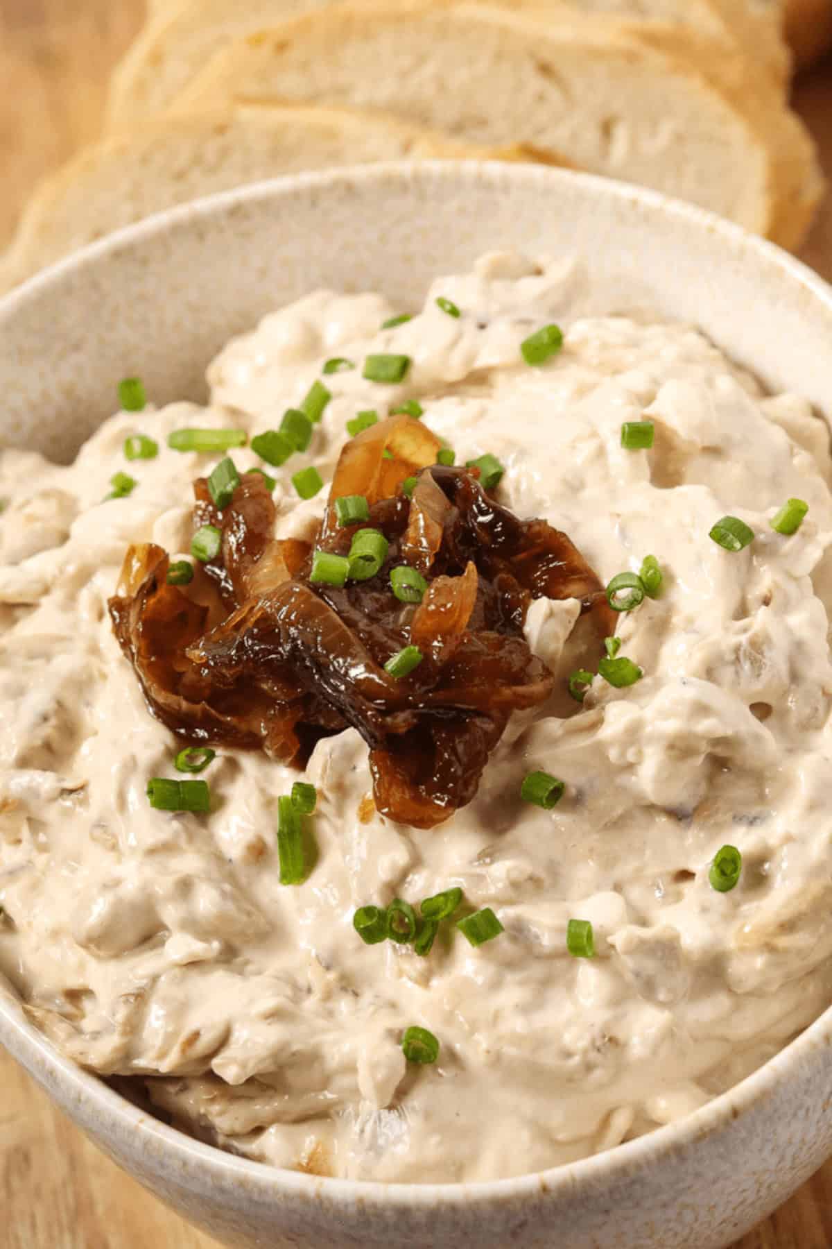 A bowl of rich caramelized onion dip is topped with chopped chives, with sliced bread in the background.