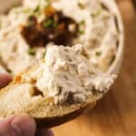 A hand holds a piece of bread topped with creamy caramelized onion dip. In the background, a bowl of dip garnished with chopped chives and sweet caramelized onions is visible.