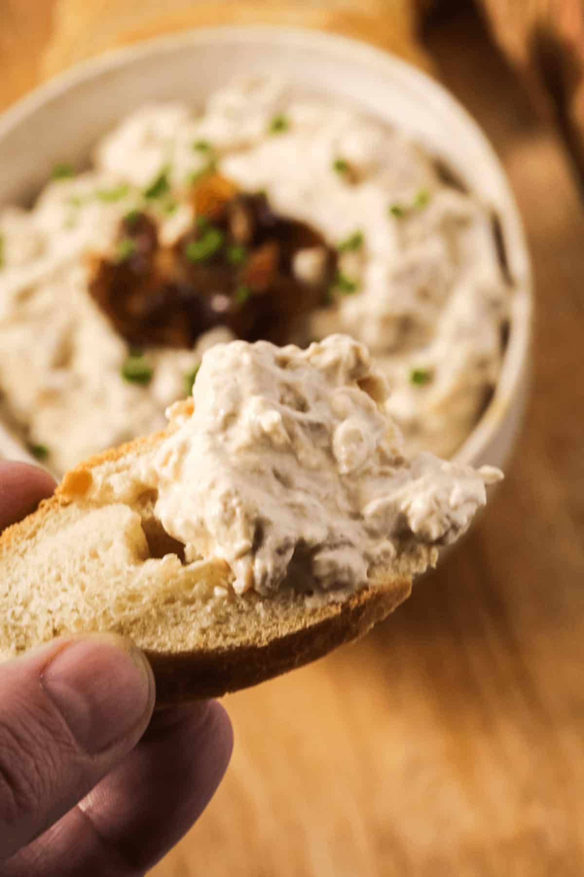 A hand holds a piece of bread topped with creamy caramelized onion dip. In the background, a bowl of dip garnished with chopped chives and sweet caramelized onions is visible.