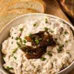 A bowl of appetizer adorned with extra caramelized onions and a sprinkle of chives, rests on a wooden board. Slices of rustic bread beckon in the background.