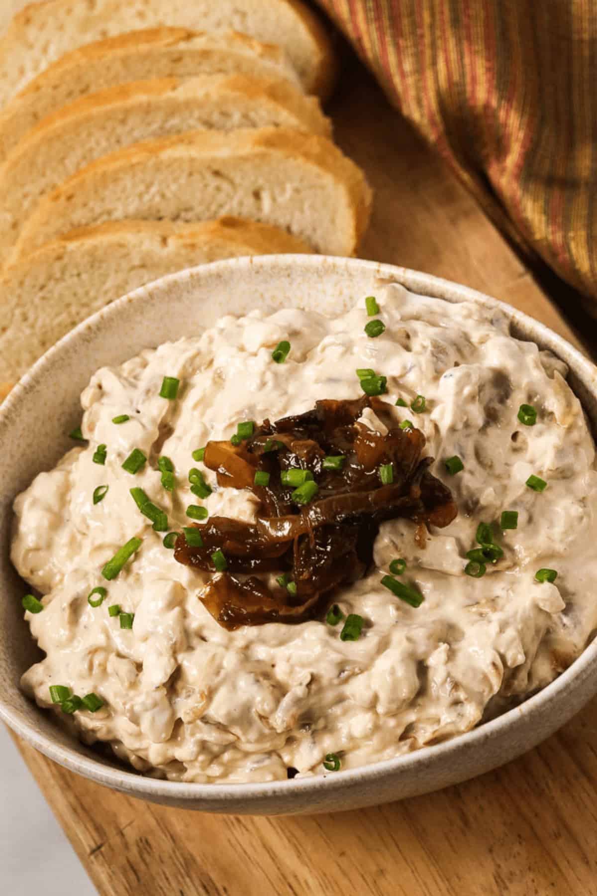 A bowl of appetizer adorned with extra caramelized onions and a sprinkle of chives, rests on a wooden board. Slices of rustic bread beckon in the background.
